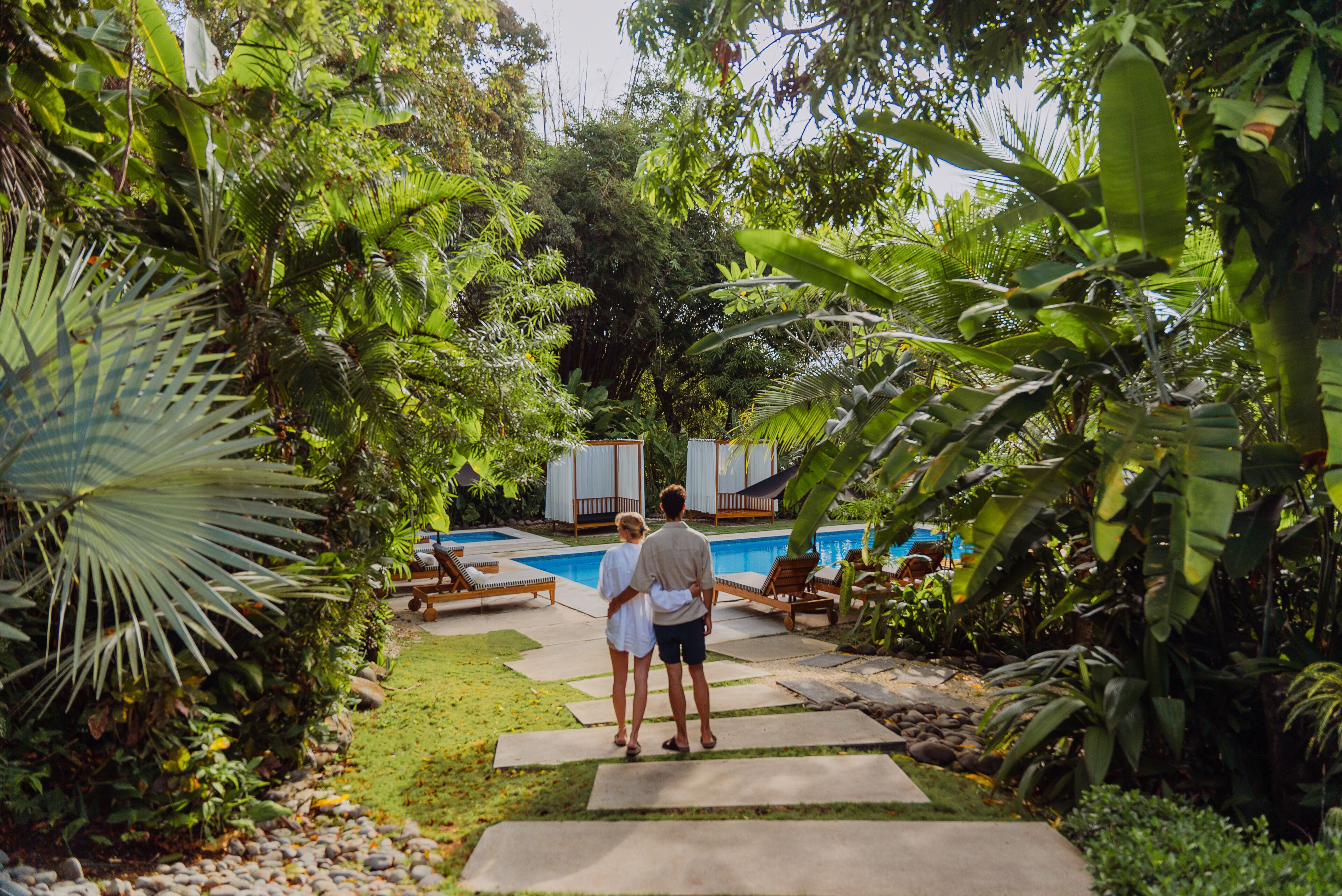 A couple walking towards a pool in a lush garden with a clear view of the pool area.