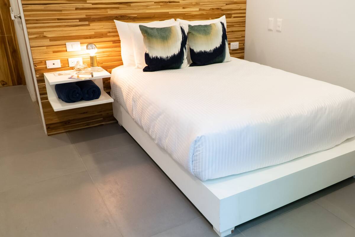 Quarto moderno com cabeceira de madeira, roupa de cama branca e mesa de cabeceira com abajur.