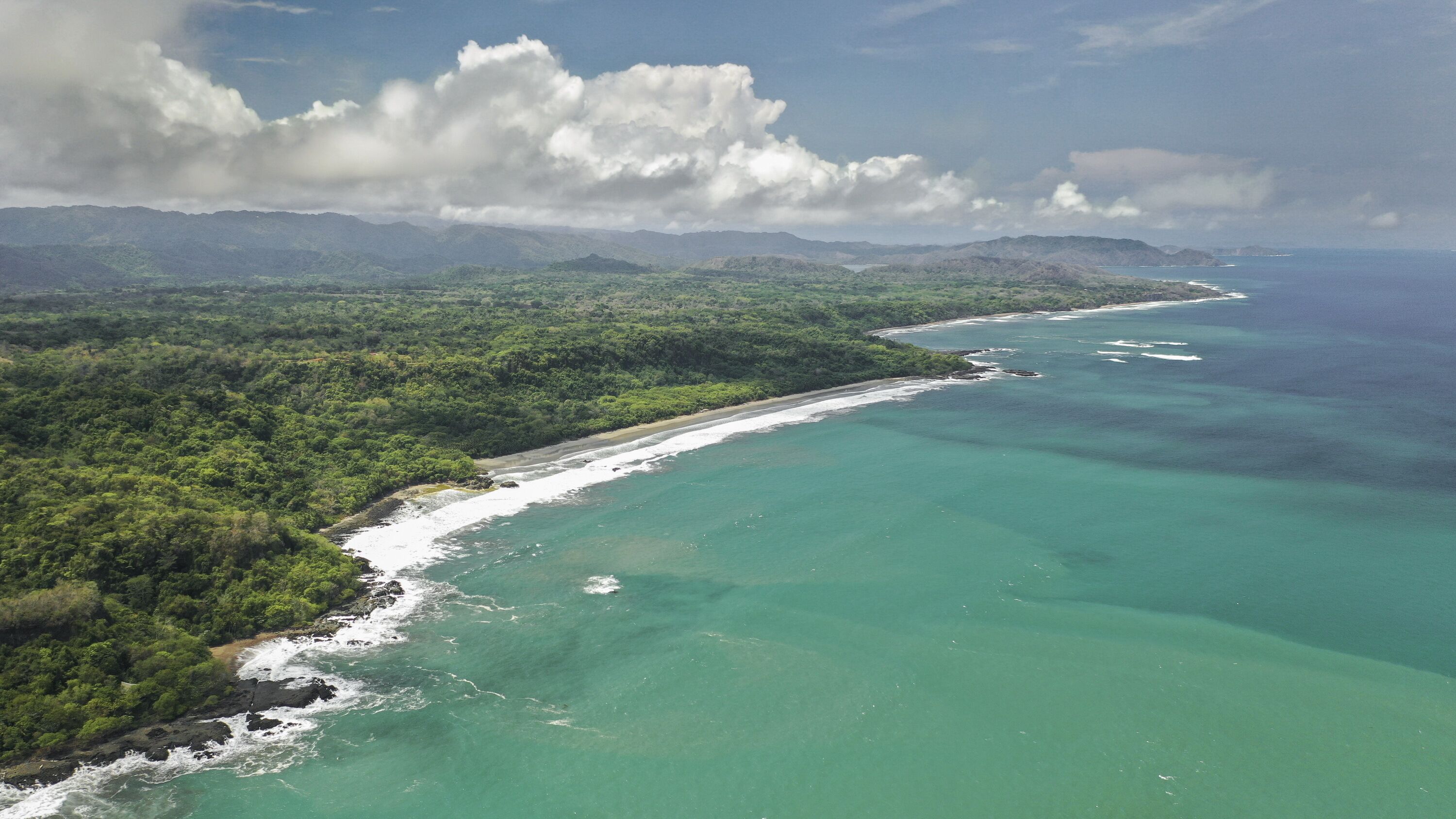 An aerial view of a coastal area with a beach, dense greenery, and mountains in the background.