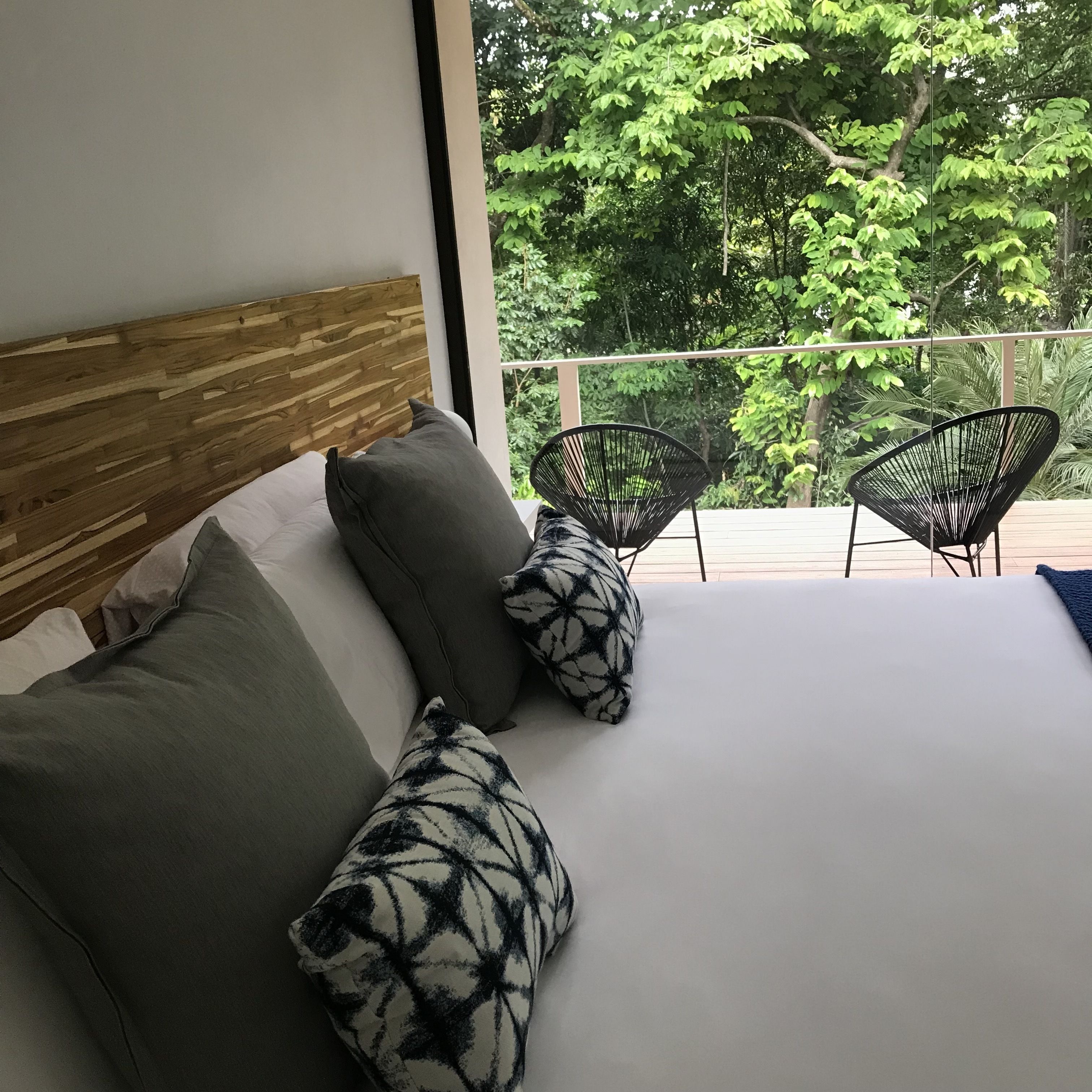 Ein Schlafzimmer mit hölzernem Kopfteil, einem großen Bett mit weißen Leinen und zwei schwarzen Stühlen auf einem Balkon mit Blick auf grüne Bäume.