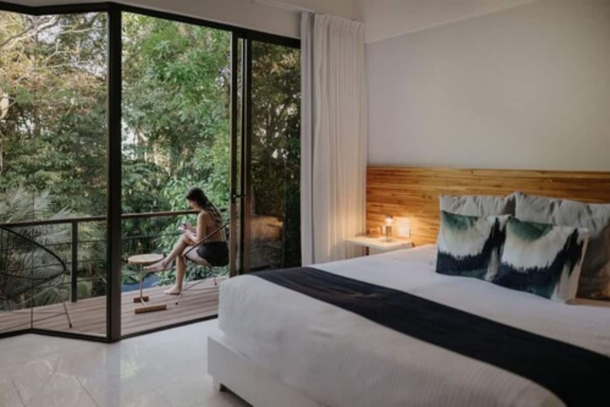 Quarto com uma cama grande, cabeceira de madeira e vista para uma varanda com uma pessoa sentada.