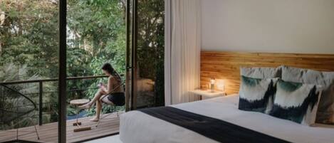 Een slaapkamer met een groot bed, een houten hoofdbord en uitzicht op een balkon met iemand die buiten zit.