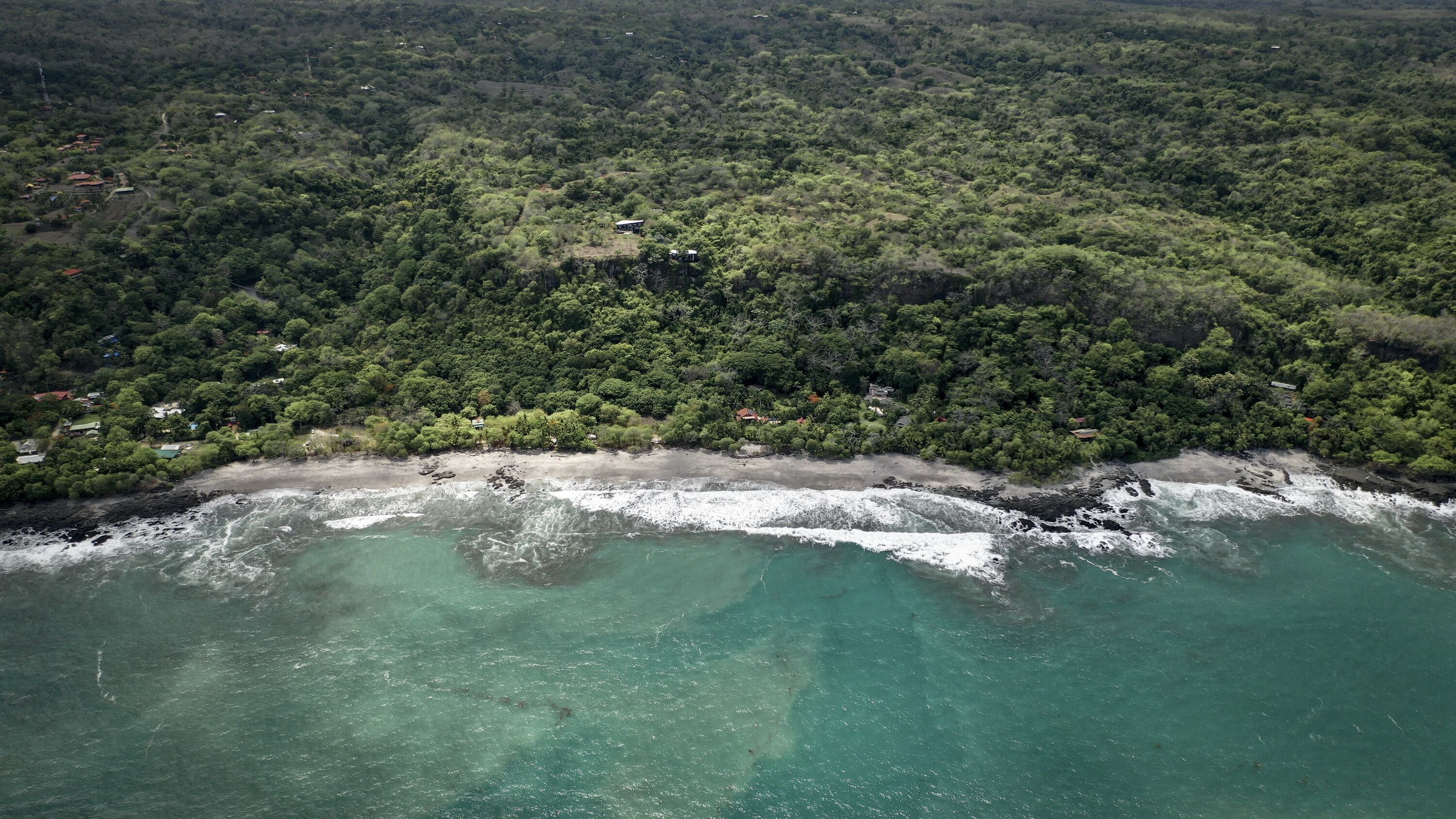 An aerial view of a coastal area with dense forest, a sandy beach, and clear turquoise waters.
