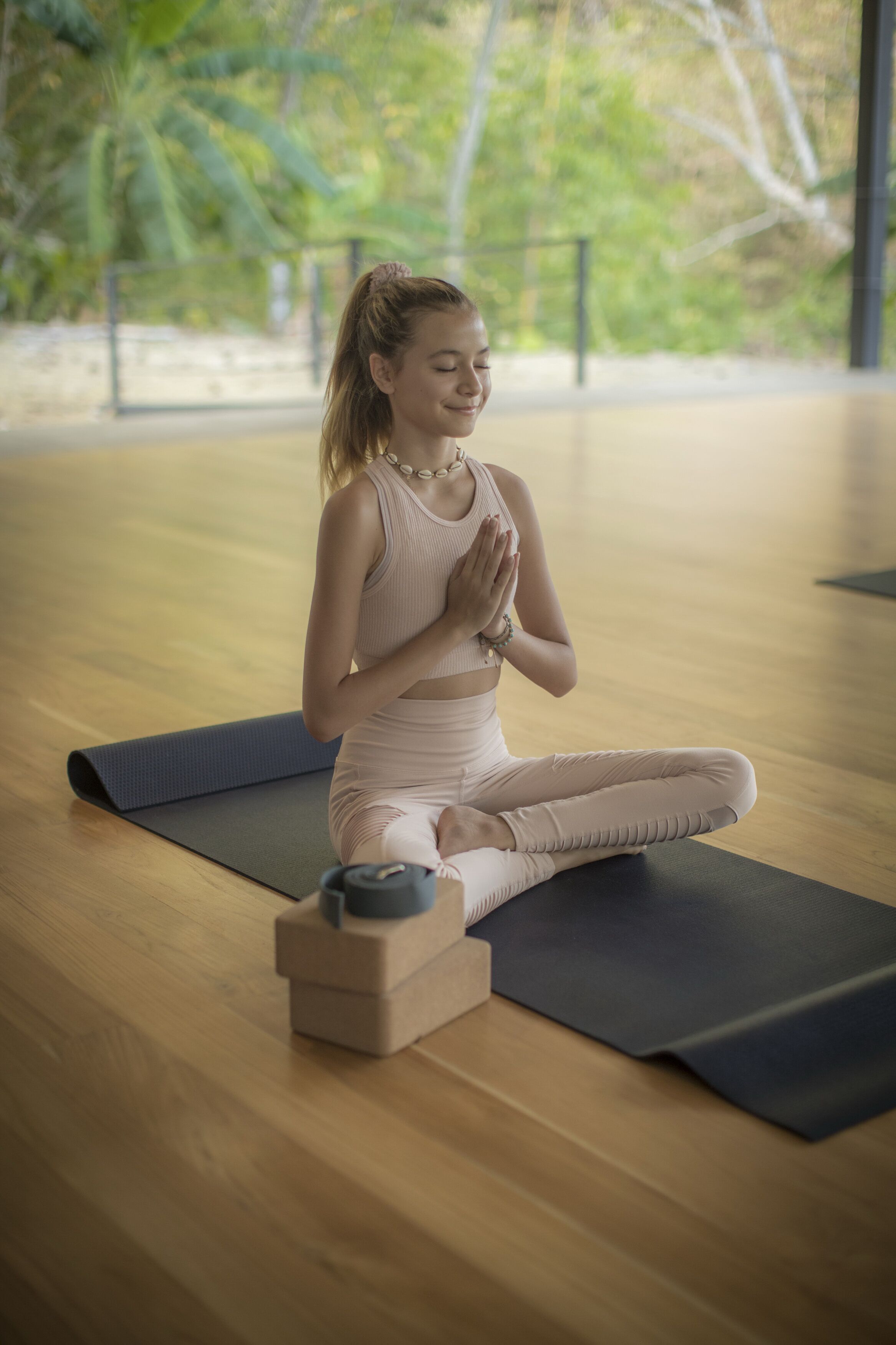 A person sitting on a yoga mat in a room with wooden flooring and a window with a view of trees.