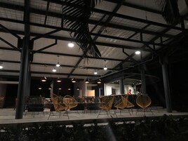 Un espacio interior con techos altos, vigas a la vista y lámparas colgantes. Varias sillas están dispuestas en filas, y se ven algunas personas al fondo.