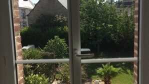 A view through a window of a garden with a brick wall, a thatched roof building, and various plants.