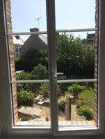 Vista pela janela de um jardim com muro de tijolos, construção de colmo e diversas plantas.