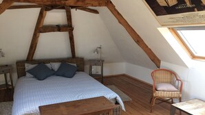 A cozy attic bedroom with a wooden ceiling, a bed with checkered bedding, a wicker chair, and a small table.