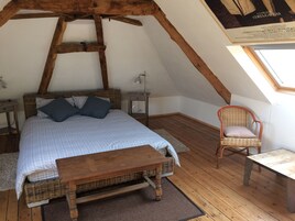 Quarto de sótão acolhedor com teto de madeira, cama com roupa de cama xadrez, uma cadeira de vime e uma pequena mesa.