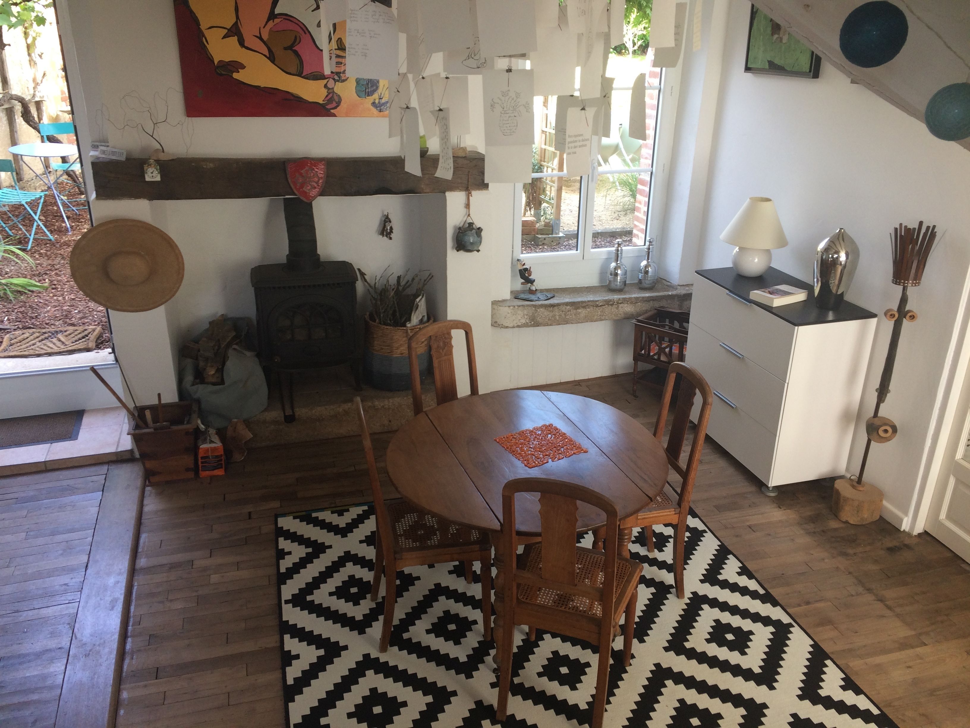 A cozy room with a wooden table, chairs, a patterned rug, a wood-burning stove, and a window with a view of the outdoors.