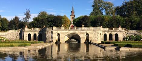A stone bridge with arches over a calm water body, surrounded by lush greenery and a distant church spire.