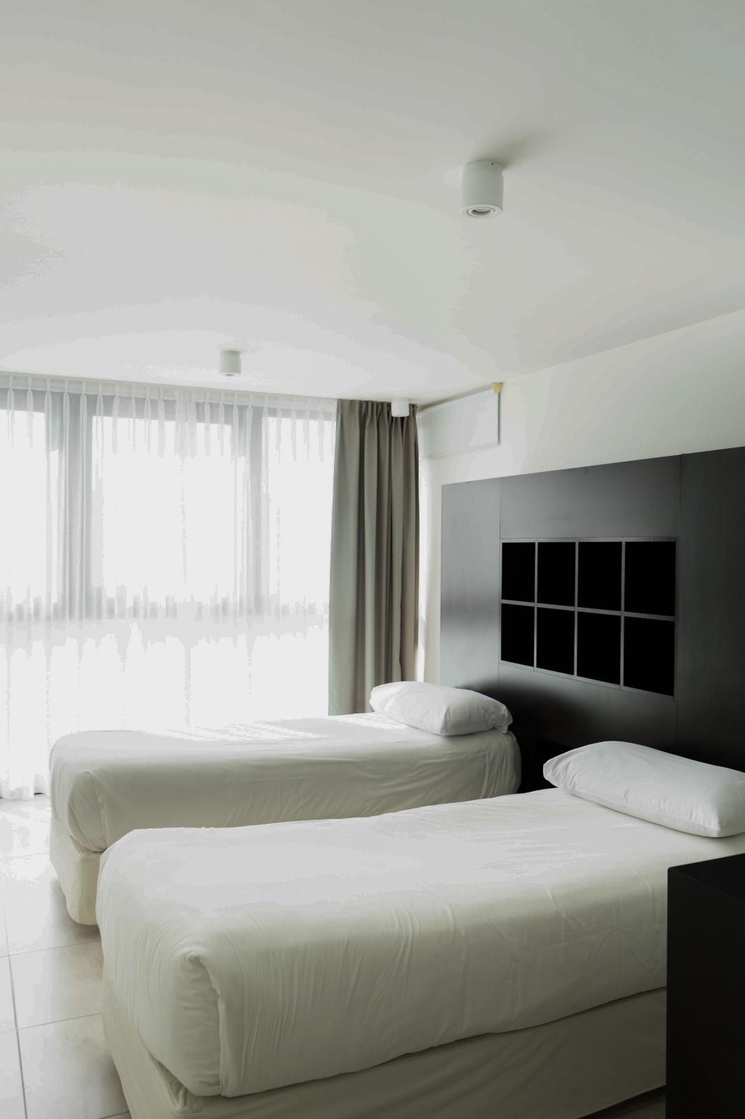 Una habitación de hotel con dos camas, un ventanal con cortinas y un cabecero moderno.