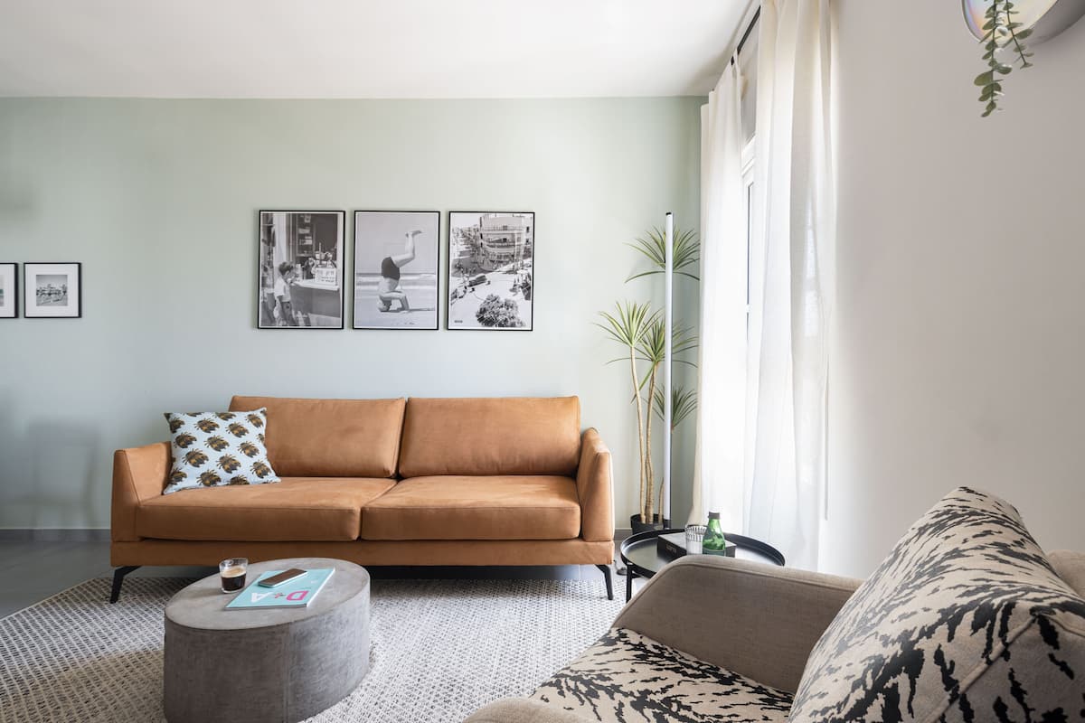 A living room with a brown sofa, a round coffee table, and a patterned armchair. There are three framed pictures on the wall and a potted plant.