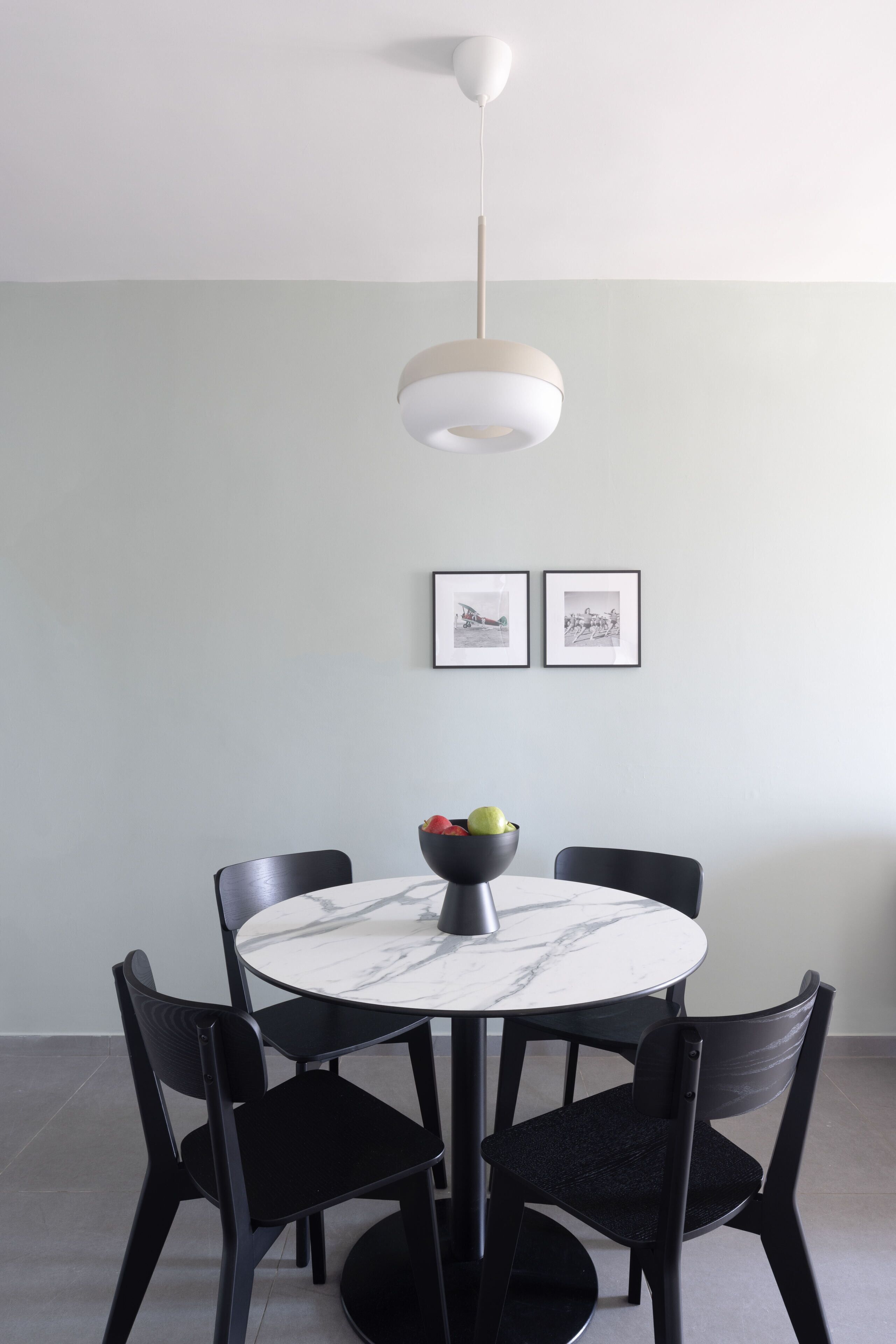 Un coin repas avec une table ronde, des chaises noires, un saladier de fruits et deux tableaux encadrés accrochés au mur.