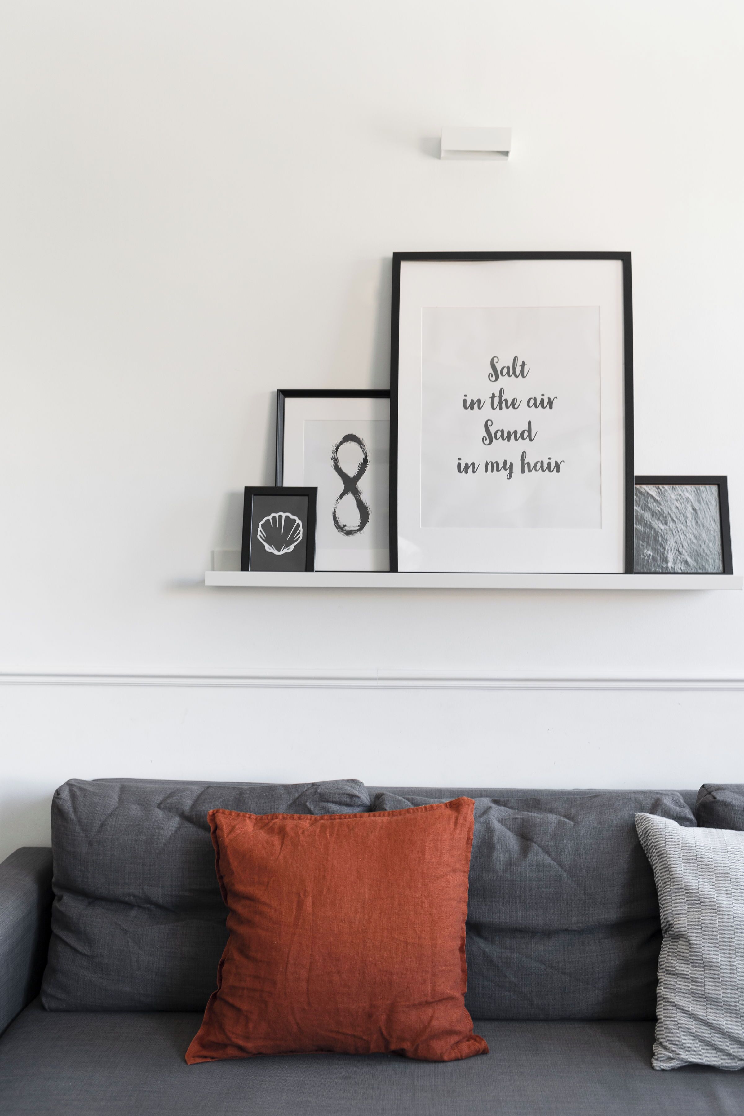 A modern living room with a grey sofa, a white shelf displaying framed artwork, and a single orange pillow.