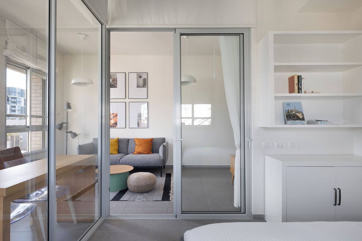 A modern, well-lit room with a glass door leading to a balcony, a sofa with cushions, a small table, and a bookshelf.