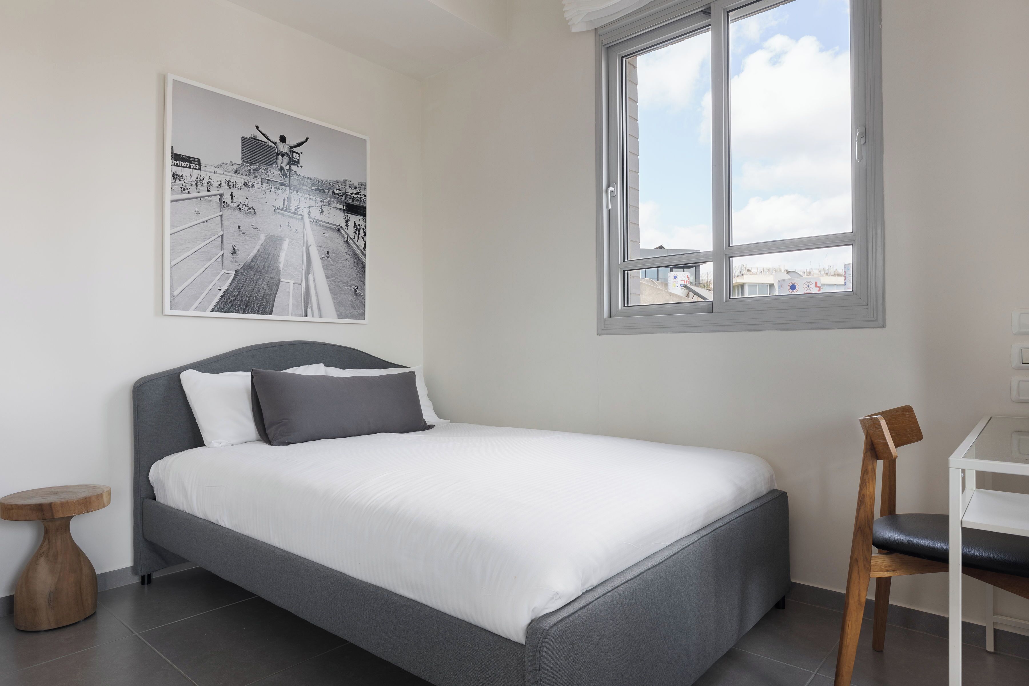 A modern bedroom with a bed, a small wooden side table, a chair, a window, and a framed picture on the wall.