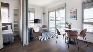 A modern, well-lit room with a bed, a seating area with a table and chairs, a TV, and large windows offering a city view. - Bauhaus Boutique by TLV2rent (Tel Aviv)