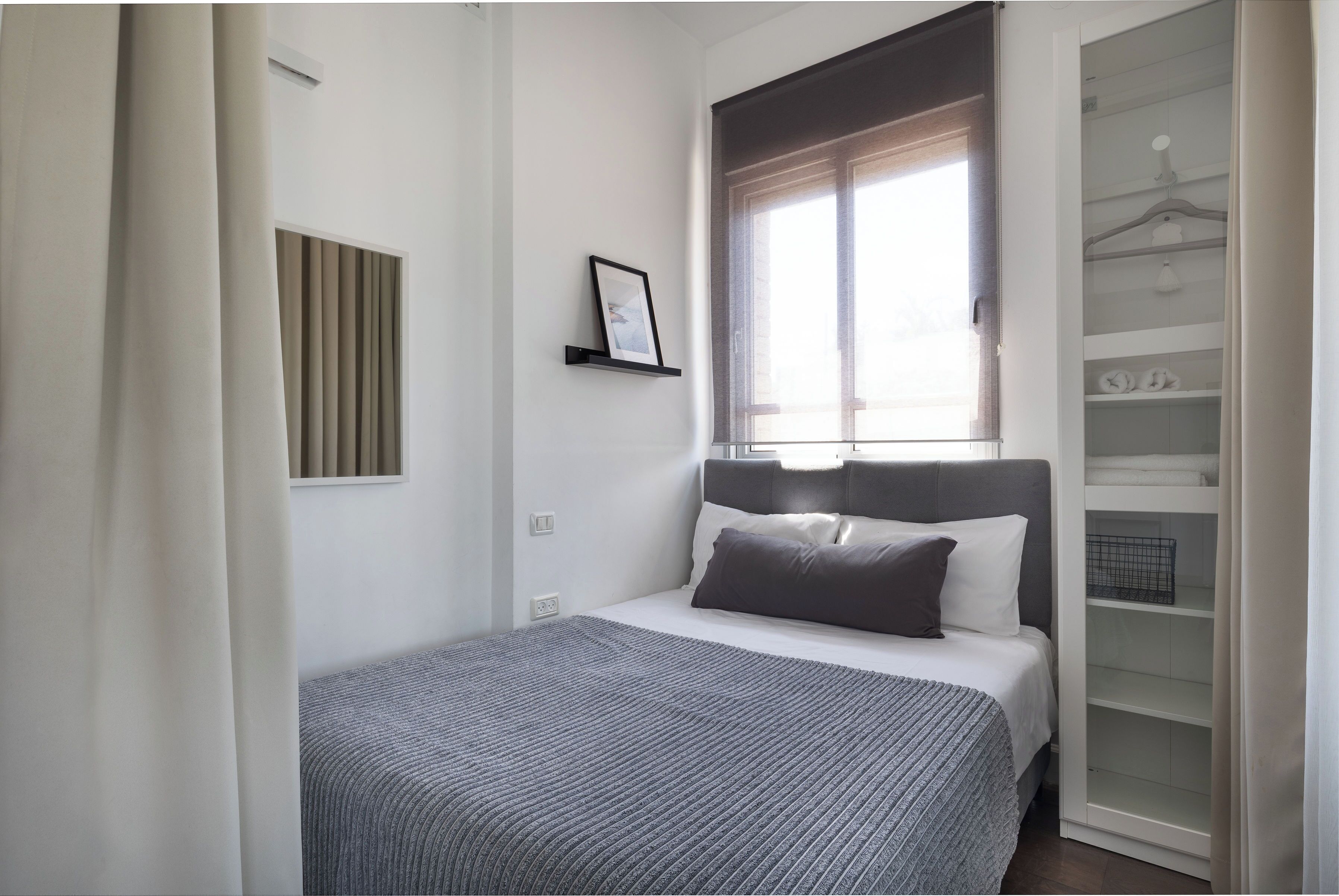 A bedroom with a bed, a window with blinds, a bedside table, and a wardrobe.