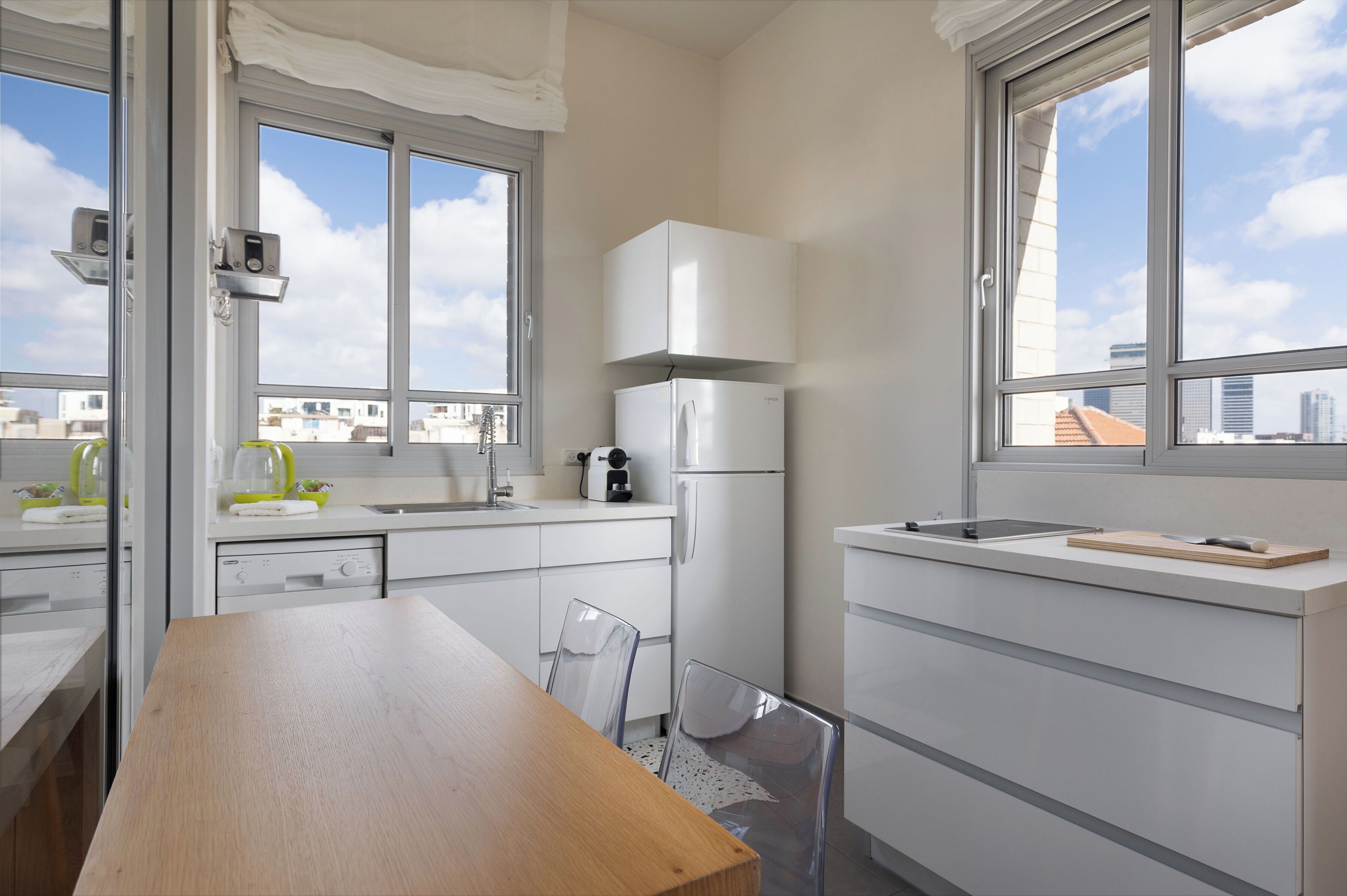 A modern kitchen with white cabinetry, a wooden dining table, and large windows offering city views.