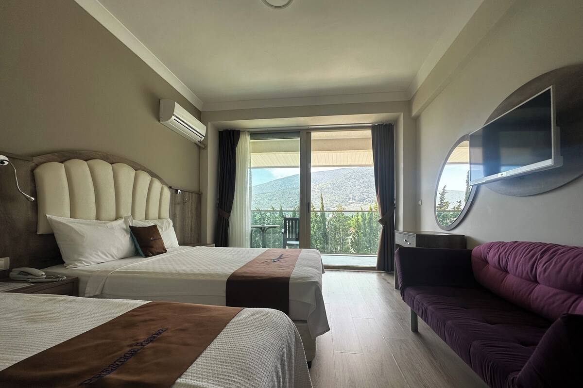 A hotel room with two beds, a sofa, a TV, and a balcony with a view of the mountains.