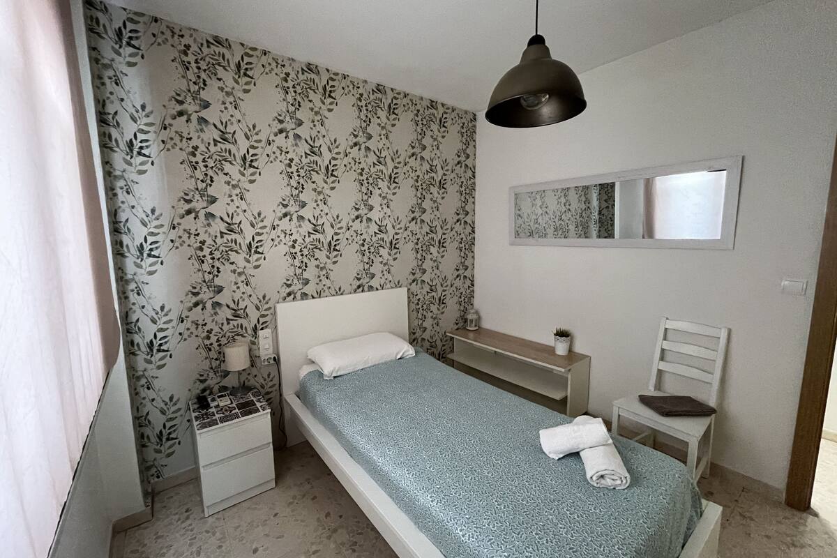 A small, neatly arranged bedroom with a single bed, a nightstand, a bedside table, a chair, and a hanging light fixture.