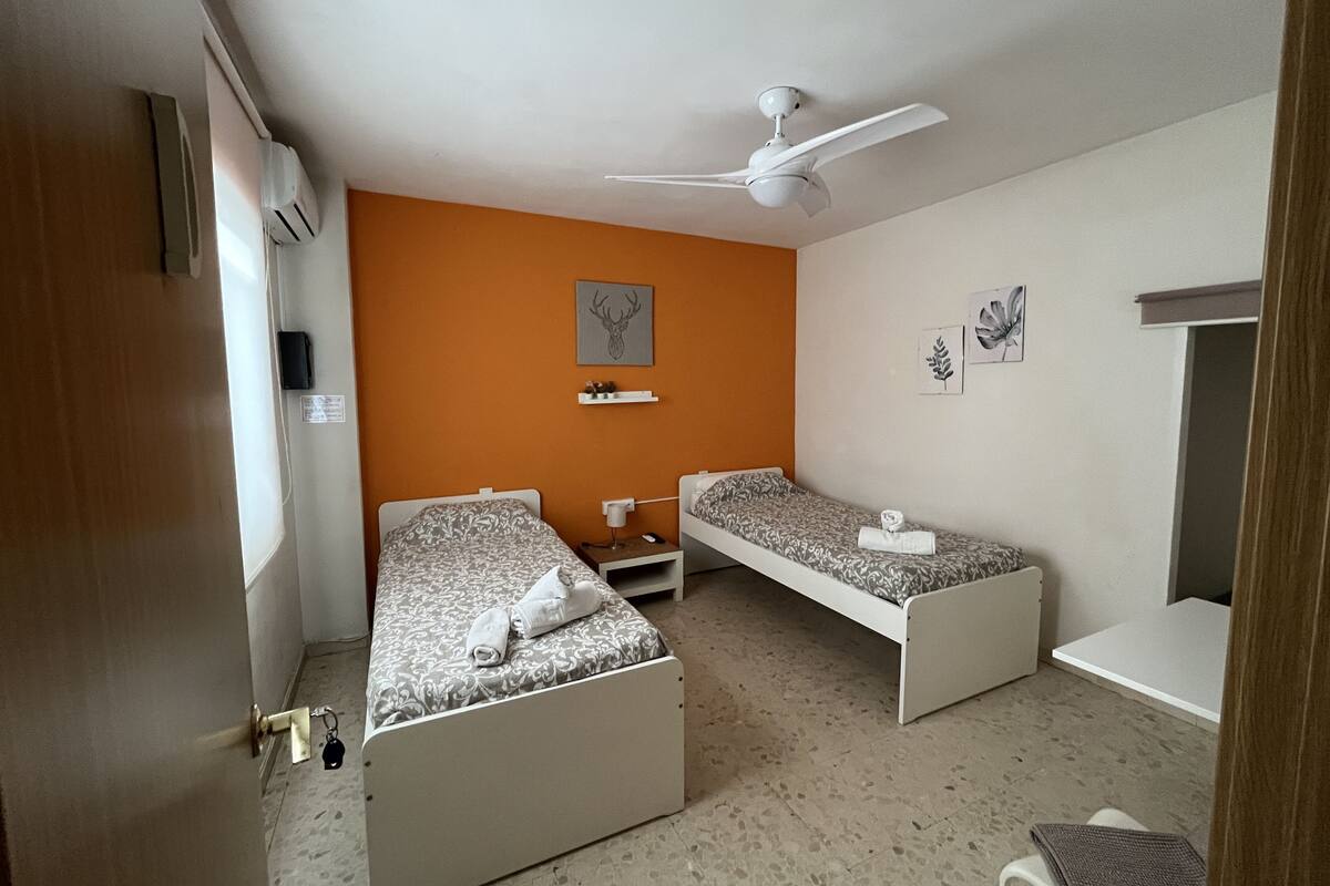 A room with two single beds, a ceiling fan, a small table, and a wall-mounted shelf.