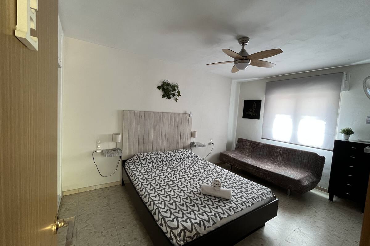 A bedroom with a bed, a ceiling fan, a bench, a window, and a small plant.