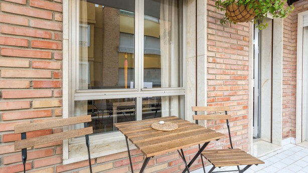 An outdoor seating area with wooden benches and a table, a hanging plant, and a brick wall. - Rafaela Guest House (Málaga)