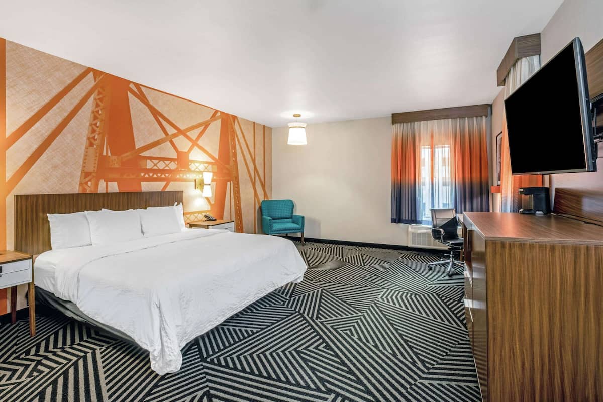 A hotel room with a large bed, a desk with a flat-screen TV, a chair, and a patterned carpet.