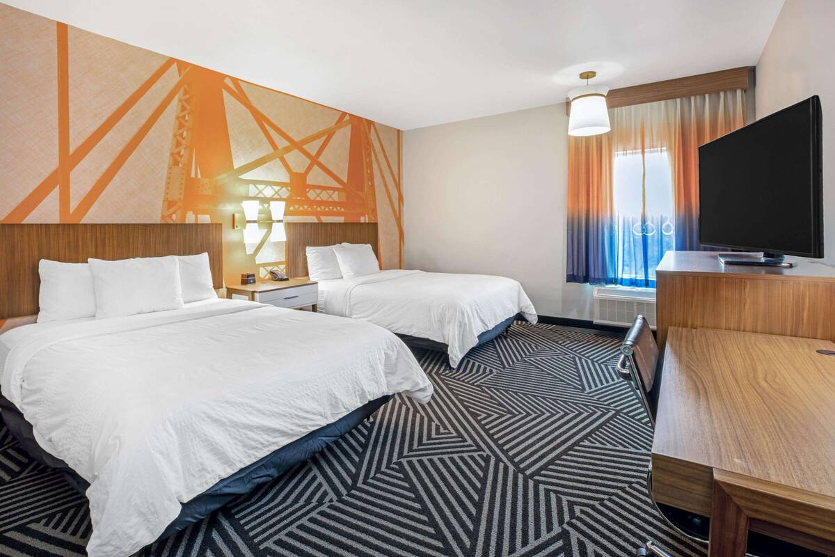 A hotel room with two beds, a desk, a TV, and a large window with curtains.