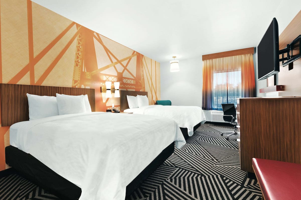 A hotel room with two beds, a desk, a TV, and a large window with curtains.