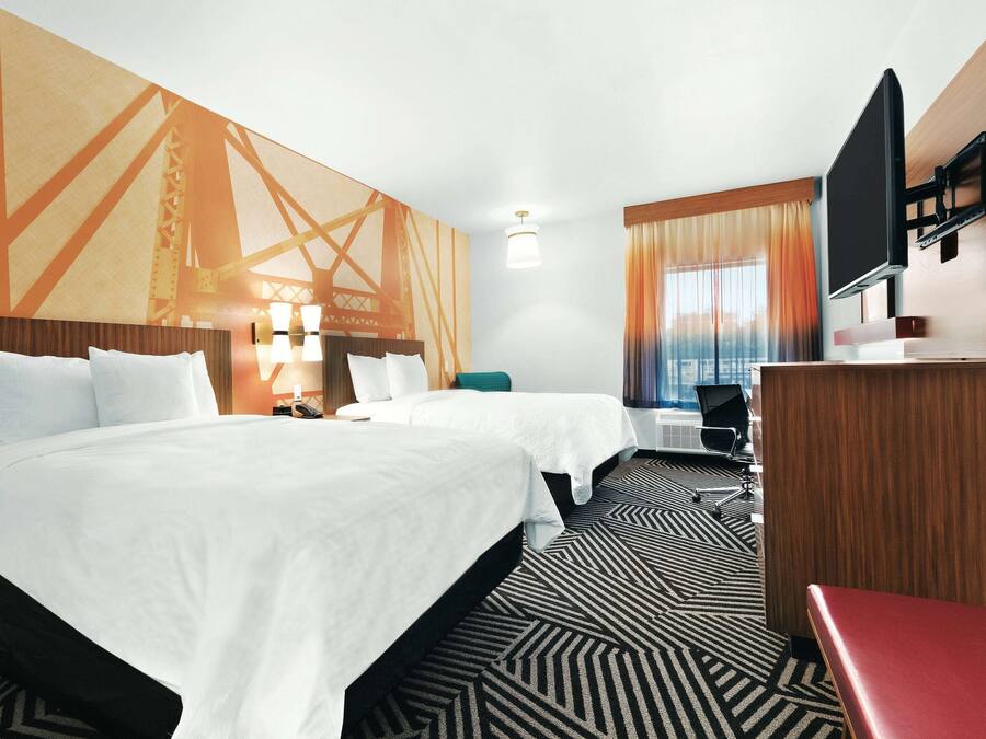 A hotel room with two beds, a desk, a TV, and a large window with curtains.