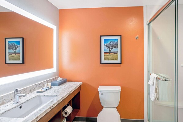 Un baño con pared naranja, un lavamanos de mármol, dos cuadros enmarcados, un inodoro y una puerta de ducha de cristal.