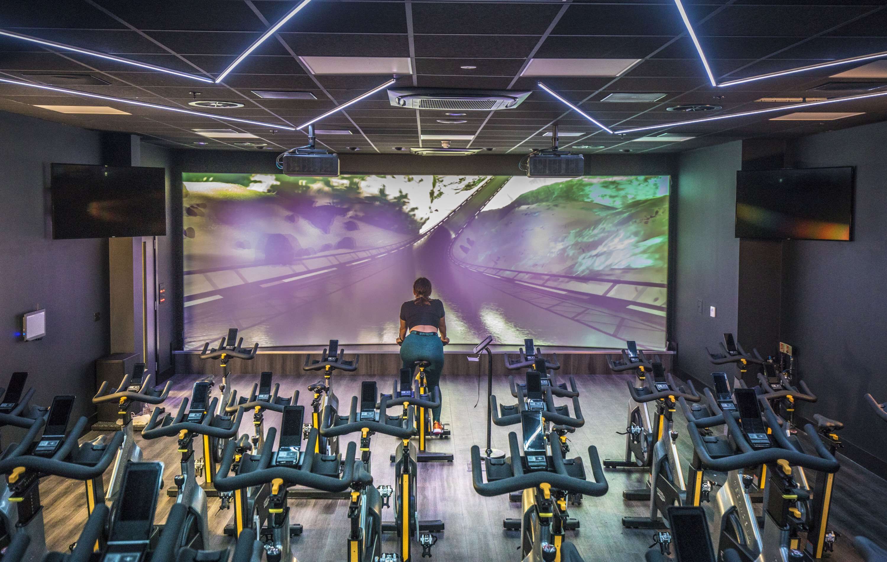 A gym with stationary bikes arranged in rows, a large screen displaying a scenic road, and a ceiling with geometric lighting.