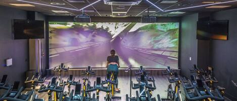A gym with stationary bikes arranged in rows, a large screen displaying a scenic road, and a ceiling with geometric lighting.