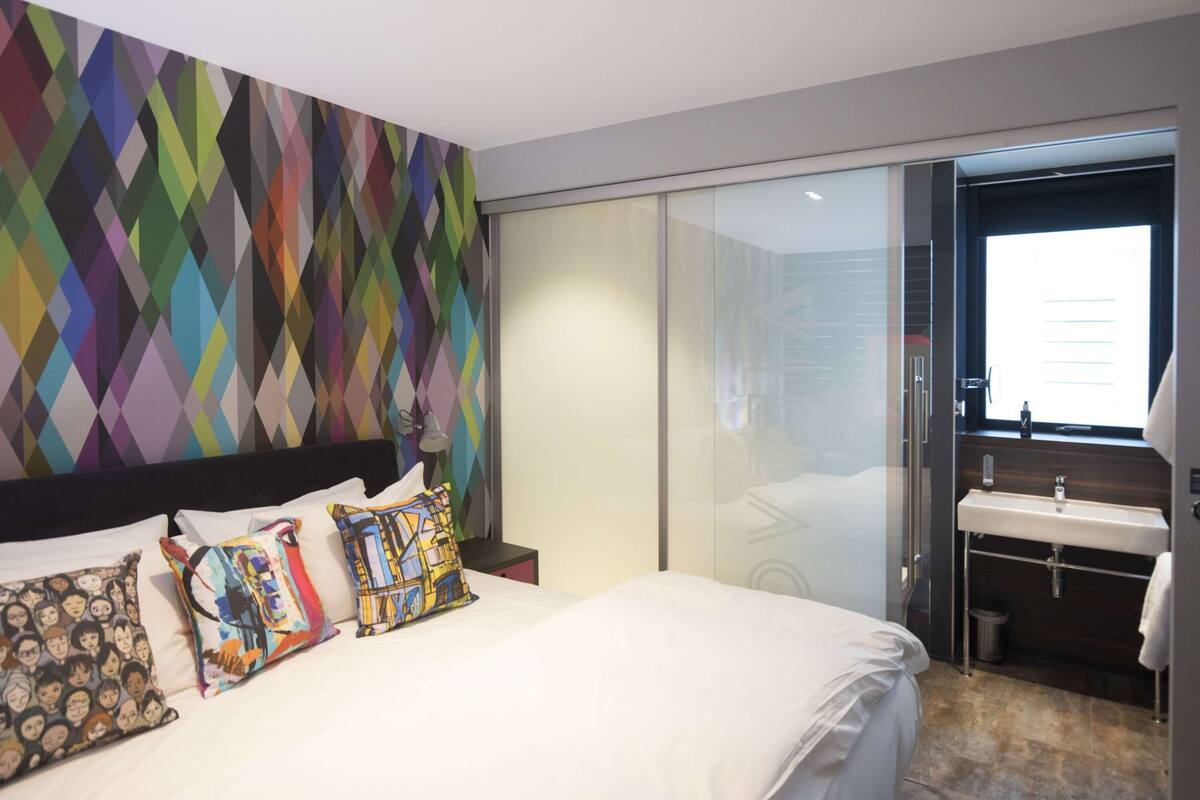 Une chambre d’hôtel moderne, dotée d’un lit, de coussins colorés, d’une grande fenêtre et d’une porte-fenêtre coulissante donnant accès à la salle de bain.