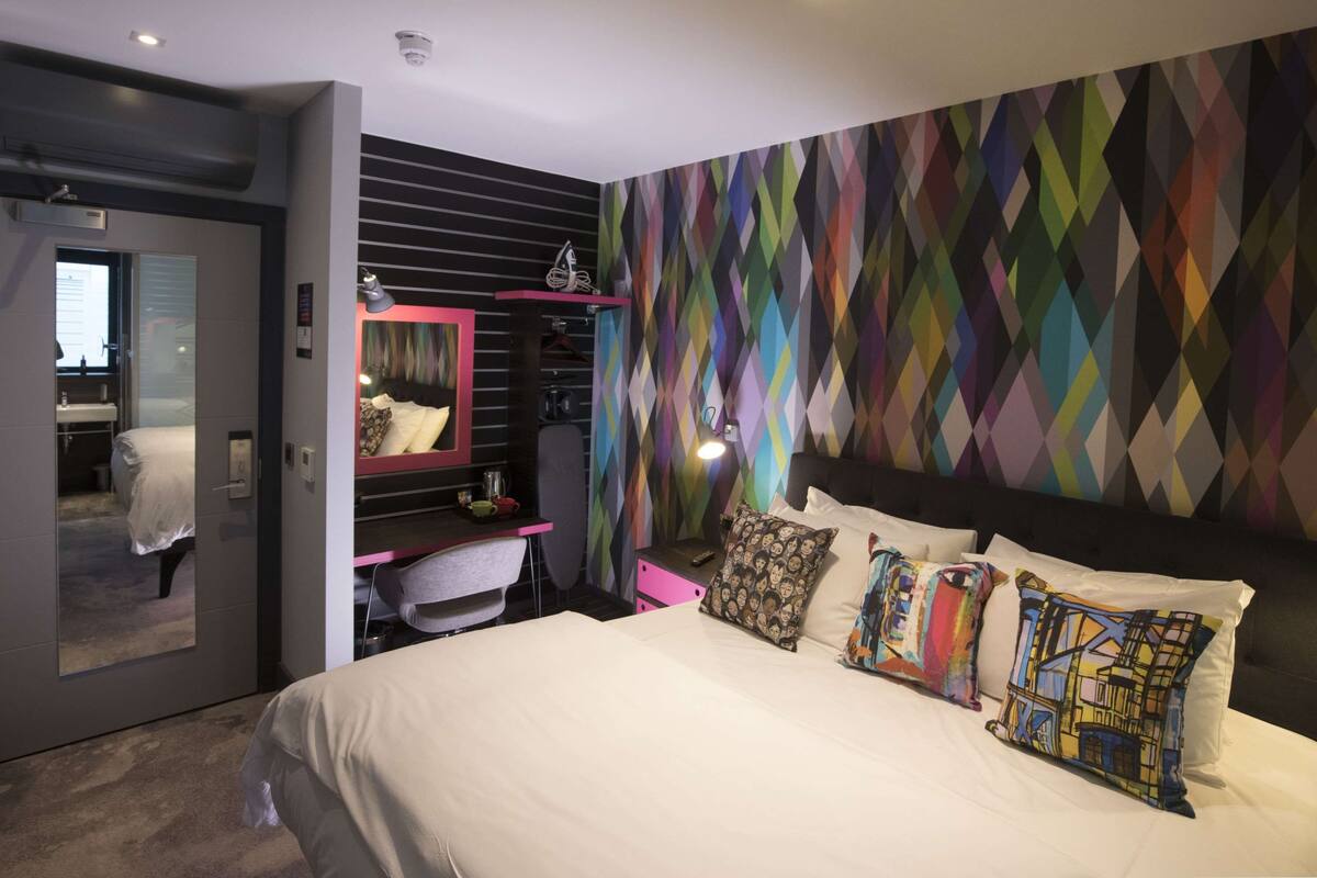 Une chambre d’hôtel moderne dotée d’un grand lit, d’un bureau avec une chaise, d’une coiffeuse et d’un mur aux motifs géométriques colorés.