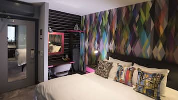 Ein modernes Hotelzimmer mit einem großen Bett, einem Schreibtisch mit Stuhl, einem Schminktisch und einer Wand mit farbigem Rautenmuster.