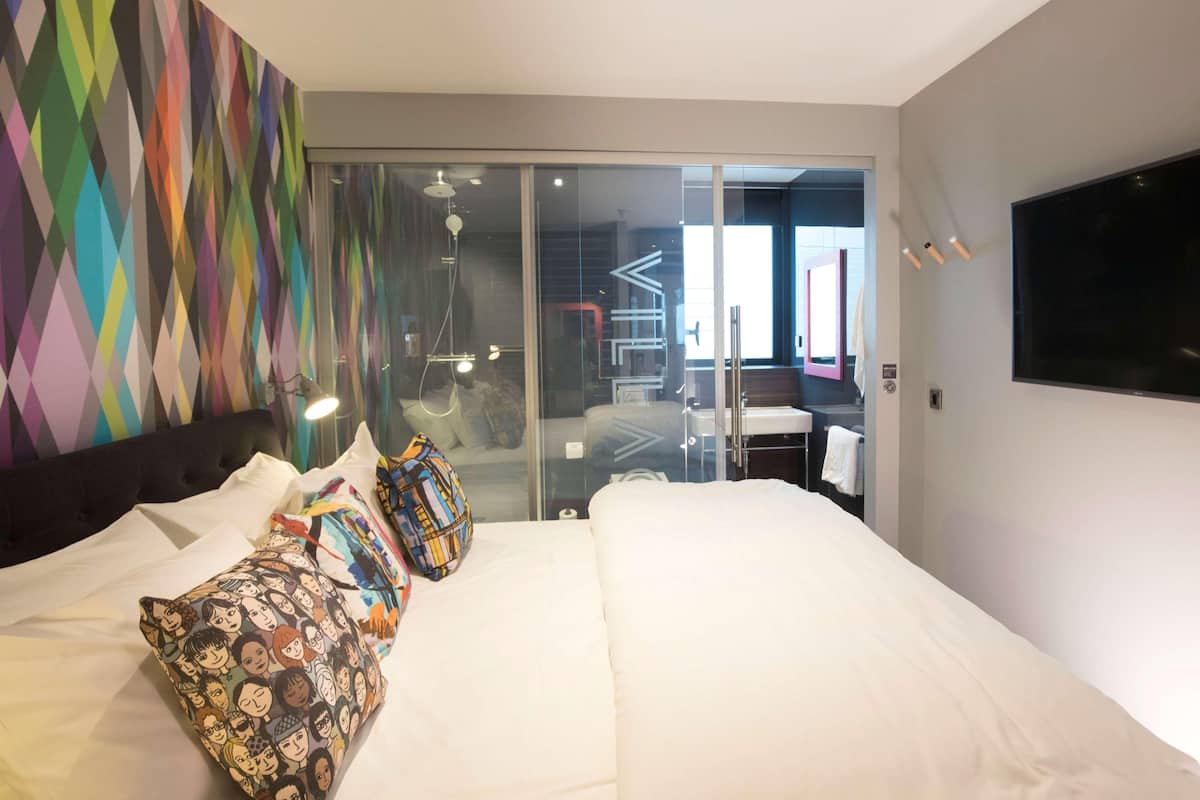 Une chambre d’hôtel moderne avec un grand lit, des œuvres d’art colorées accrochées au mur, une porte en verre donnant accès à la salle de bain et un téléviseur à écran plat fixé au mur.