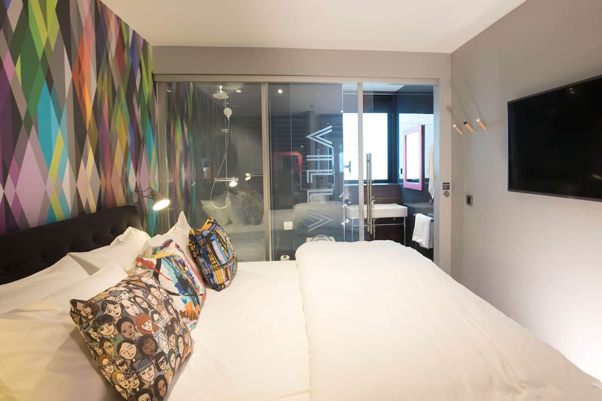 Une chambre d’hôtel moderne avec un grand lit, des oreillers colorés, une télévision et une porte-fenêtre coulissante donnant sur la salle de bain.