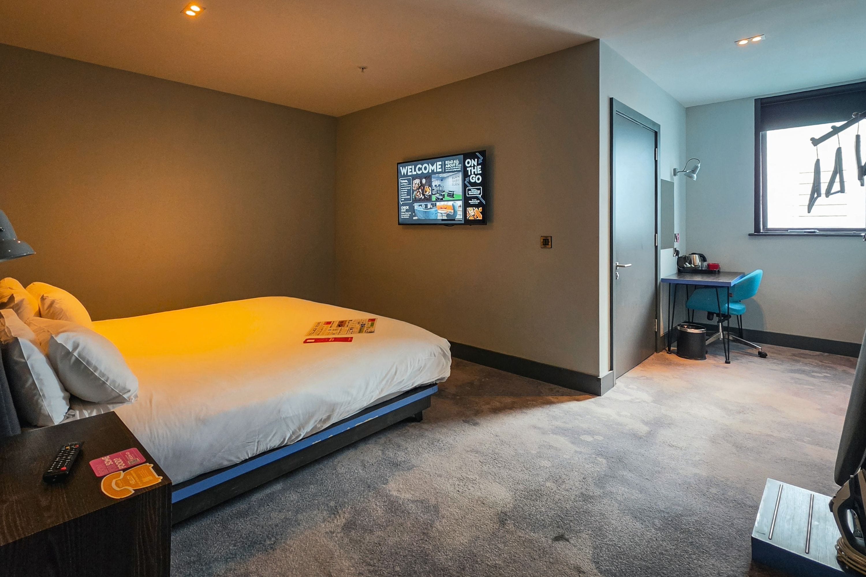 A hotel room with a bed, a desk, a chair, a desk lamp, a wall-mounted TV, and a door.