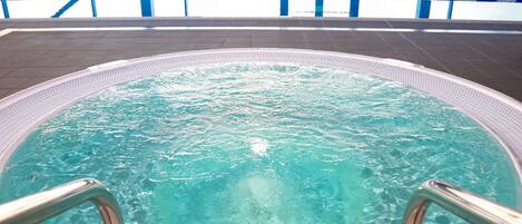A hot tub with metal railings, surrounded by a tiled deck and a pool with lane markers.