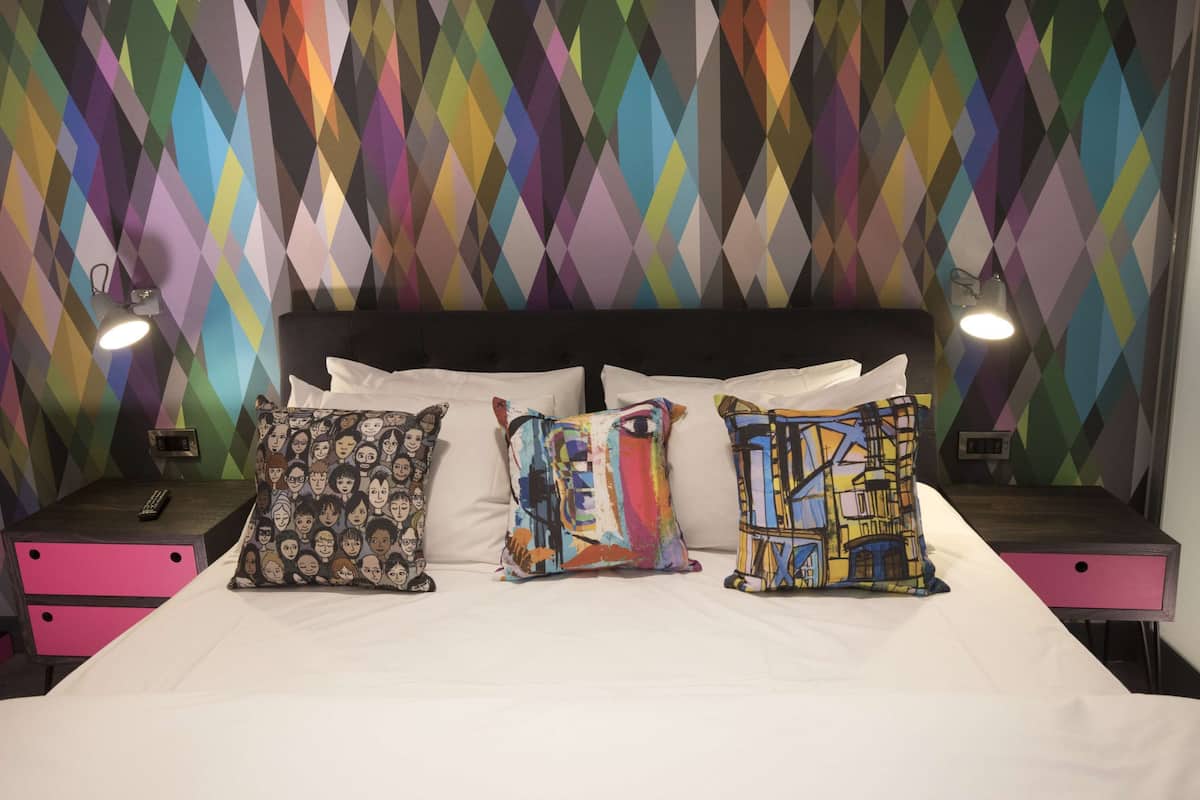Un lit avec du linge de lit blanc et trois coussins colorés, une tête de lit noire et deux tables de chevet avec des lampes.