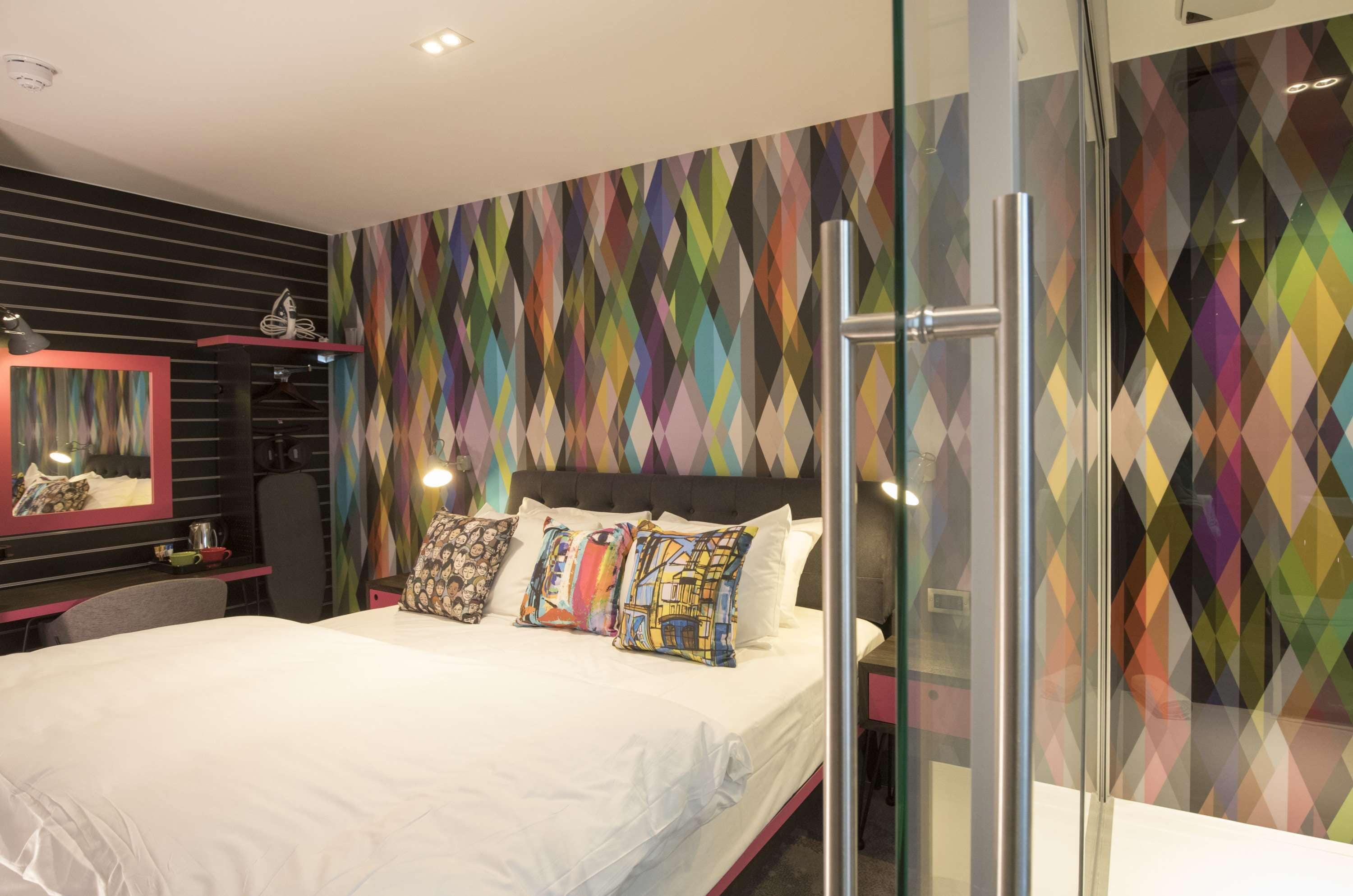 A modern hotel room with a large bed, a glass shower enclosure, and a colorful geometric-patterned wall.