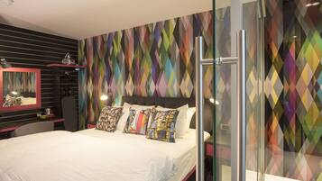 A modern hotel room with a large bed, a glass shower enclosure, and a colorful geometric-patterned wall.