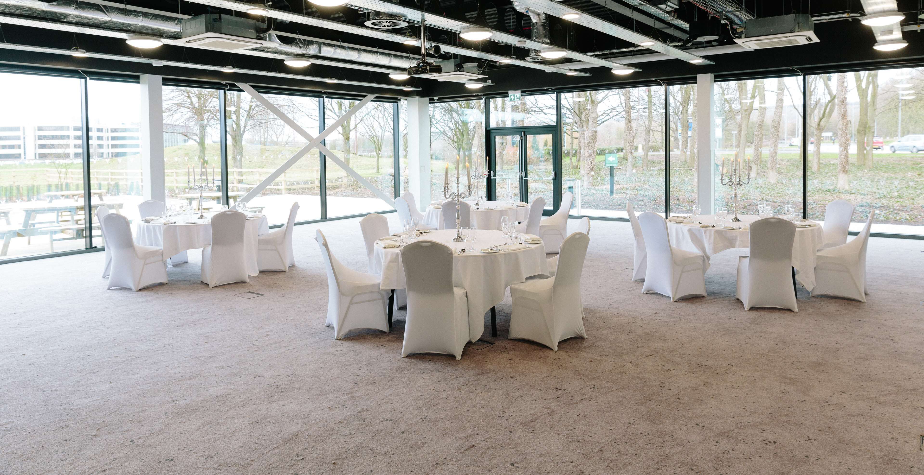 A spacious indoor venue with large windows, white tables and chairs, and a high ceiling with exposed beams.