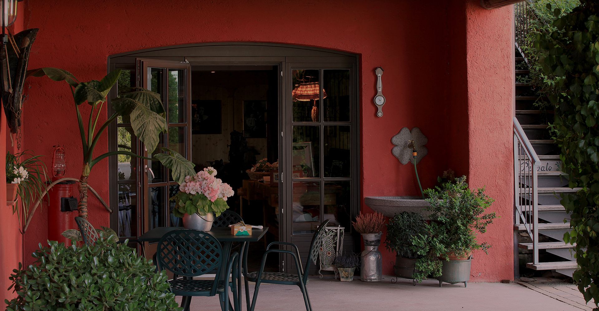 A red building with a curved entrance, a small table with chairs, and various potted plants.