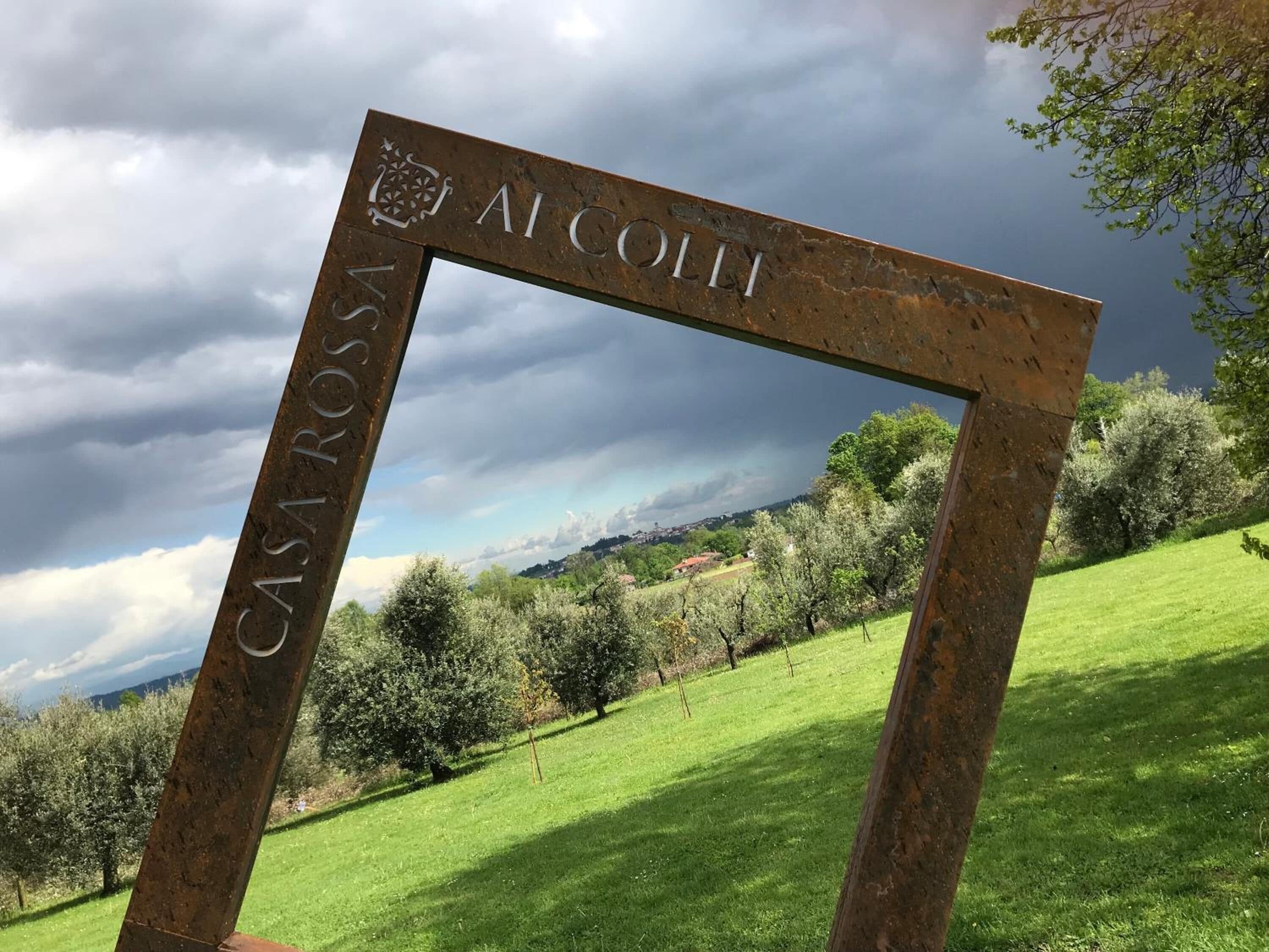 Une enseigne en métal au style rustique, portant les inscriptions « CASA ROSSA » et « ALCOH », est placée sur un fond de paysage verdoyant et luxuriant, sous un ciel nuageux.