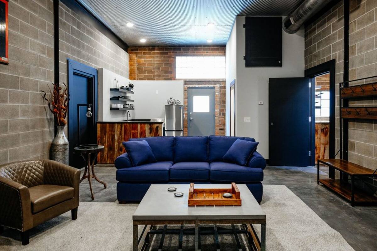 A modern living room with a blue sofa, a coffee table, and a brown leather armchair. There is a kitchen area with a refrigerator and a sink in the background.